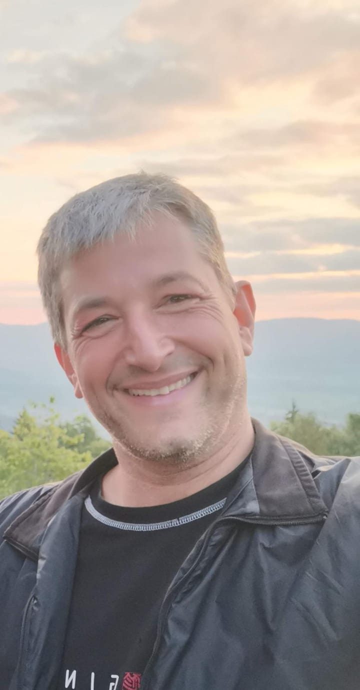 Portrait von Markus Loss: kurze graue Haare, offenes Lächeln, dunkle Jacke, im Hintergrund Abendhimmel über bewaldeten Hügeln.