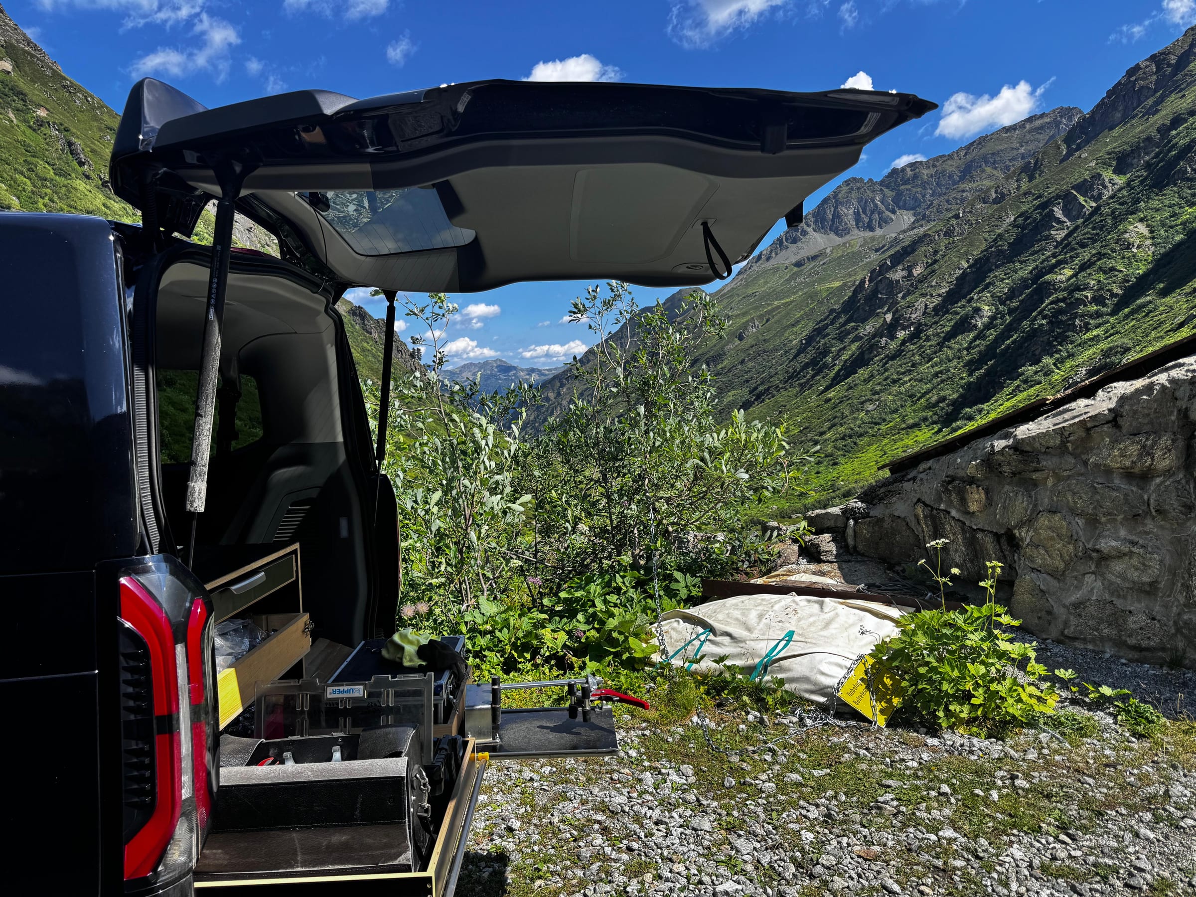 Schwarzer Servicebus mit geöffneter Heckklappe und ausgezogener Werkbank vor einem grünen Bergtal in Vorarlberg.