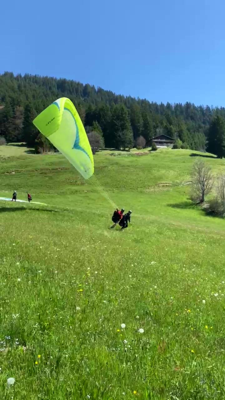 Tandemstart auf einer Wiese: Der Schirm steht, Pilot und Passagier laufen an – dahinter Wald und blauer Himmel.