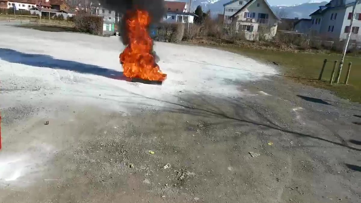 Standbild: Brennende Feuerschale auf einem Übungsplatz, davor Löschschaum auf dem Boden, dahinter Wohnhäuser