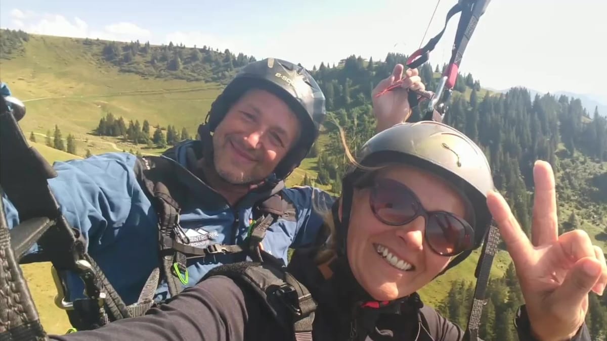 Standbild: Pilot und lachende Passagierin mit Helm im Flug, im Hintergrund grüne Berge