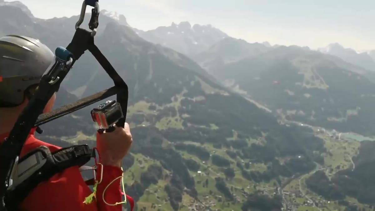 Standbild: Passagier in roter Jacke fotografiert aus dem Tandem, unter ihm das Tal mit Dörfern und die Bergkette