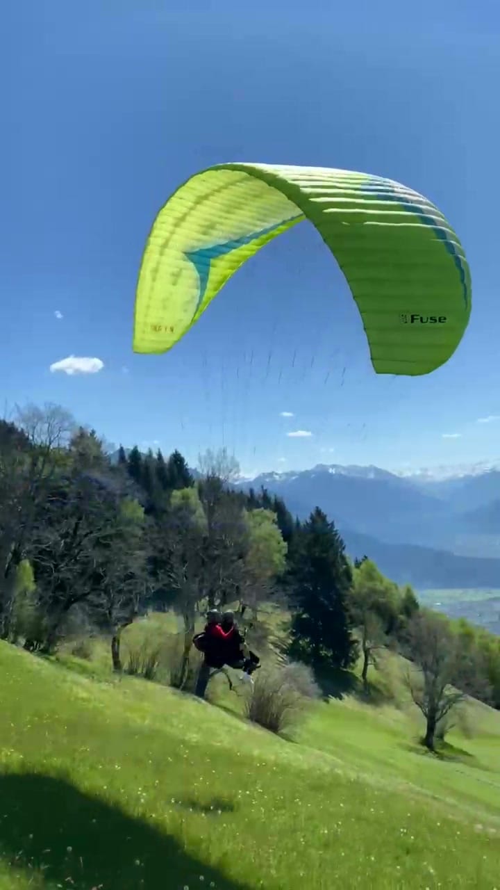 Standbild: Tandem kurz nach dem Abheben, der gelbgrüne Schirm über dem Hang, dahinter Wald und Berge